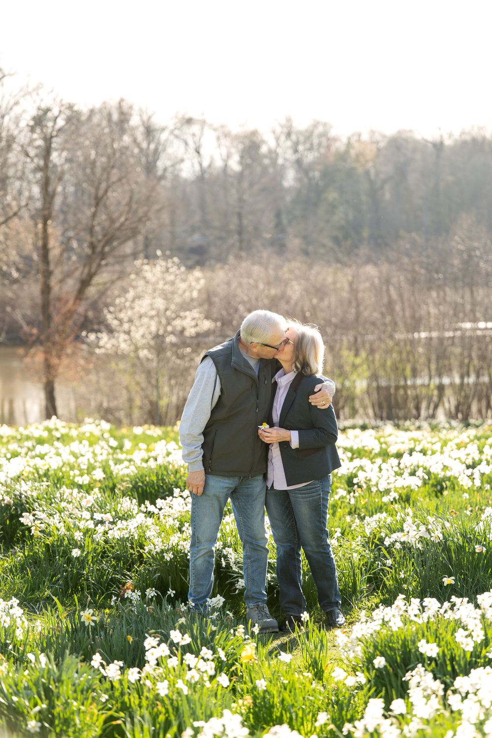 An older couple shares a tender kiss while standing together in a bright spring daffodil field.