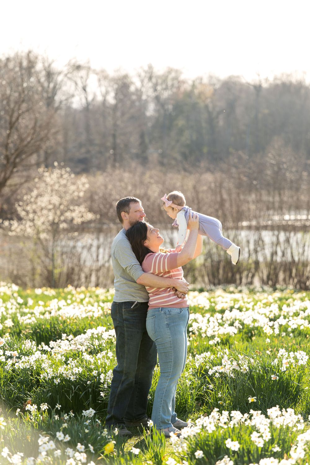 Family with baby in daffodil field, parents lifting child in sunny spring meadow near water.