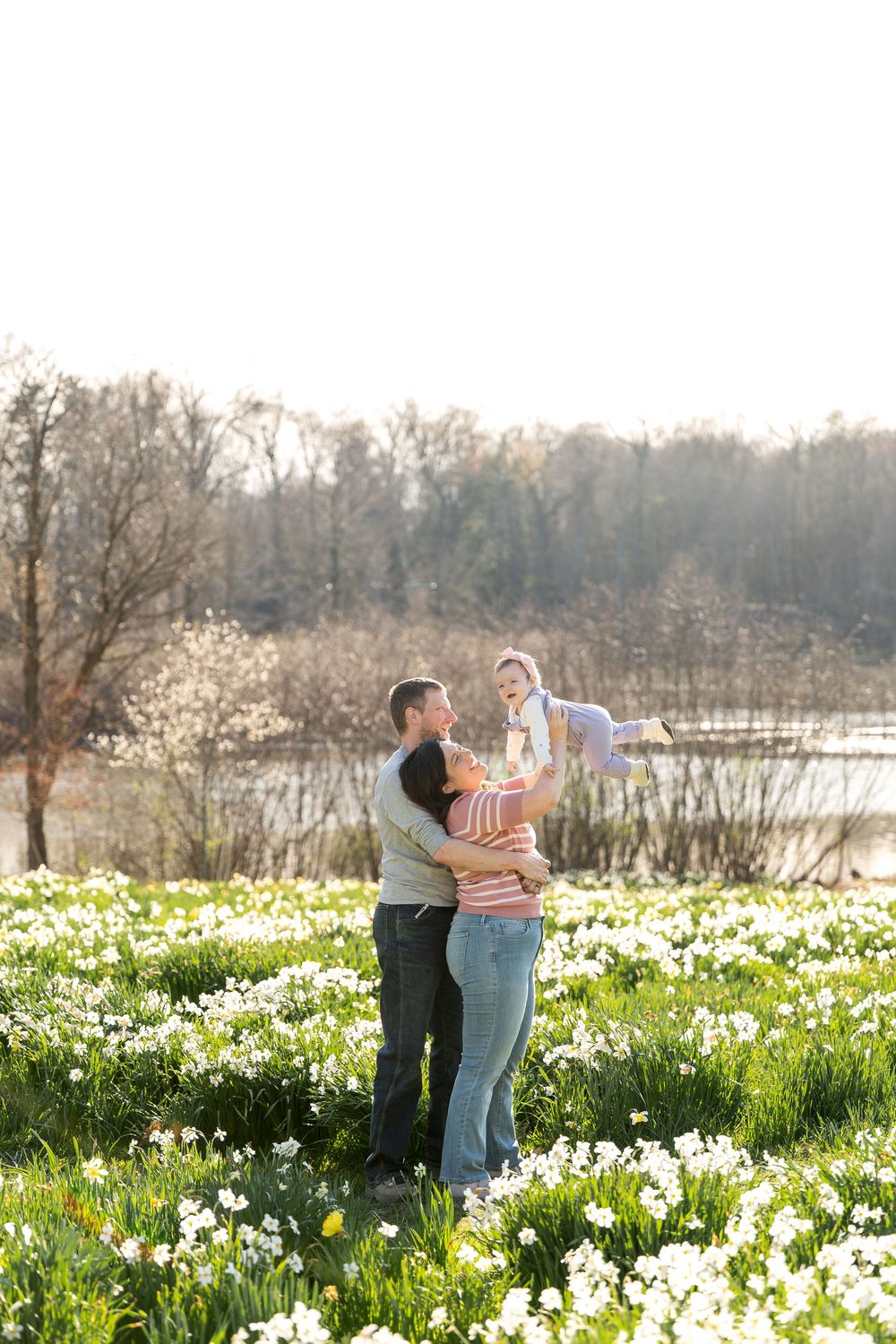Happy family standing in blooming daffodil field, mother lifting baby while father embraces her.