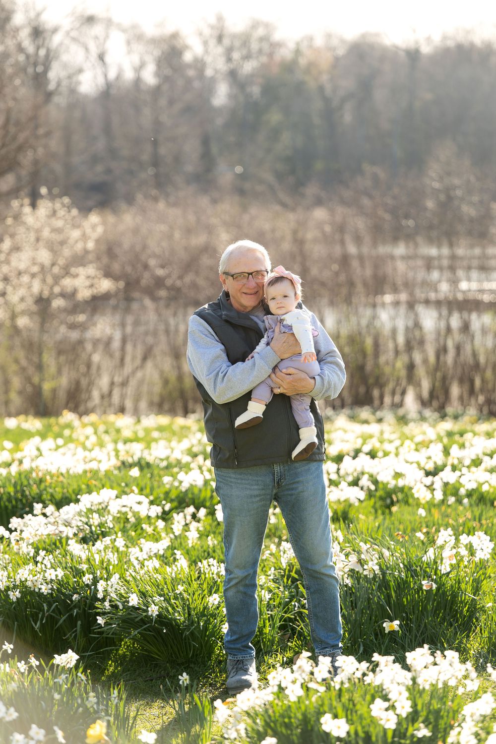 Grandfather holding baby in sunlit daffodil field, smiling at camera on a spring day.