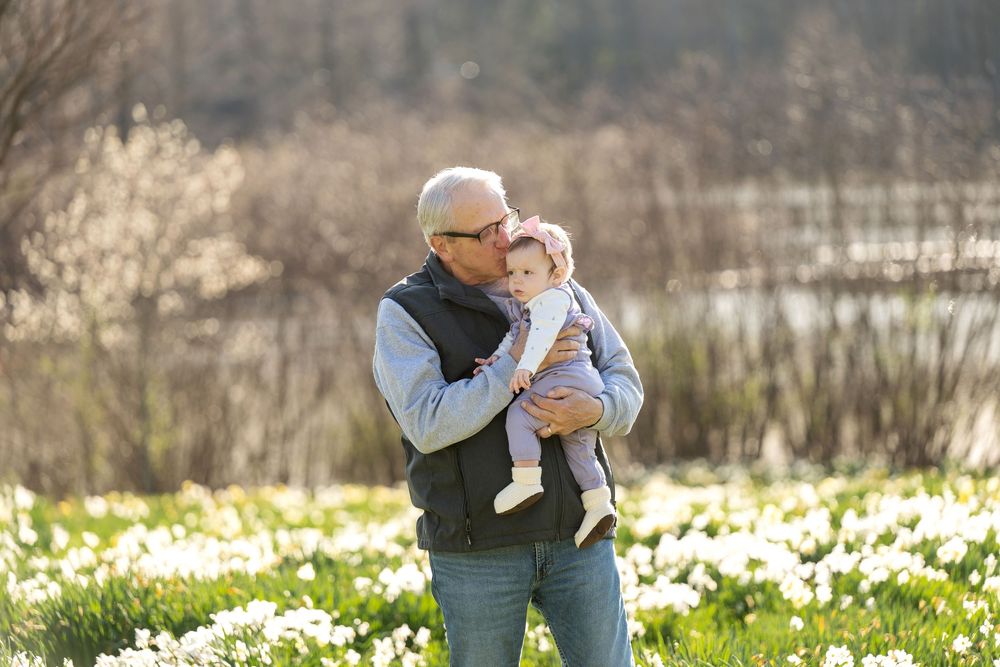 Senior man kissing baby in white daffodil meadow, sharing a tender moment outdoors.