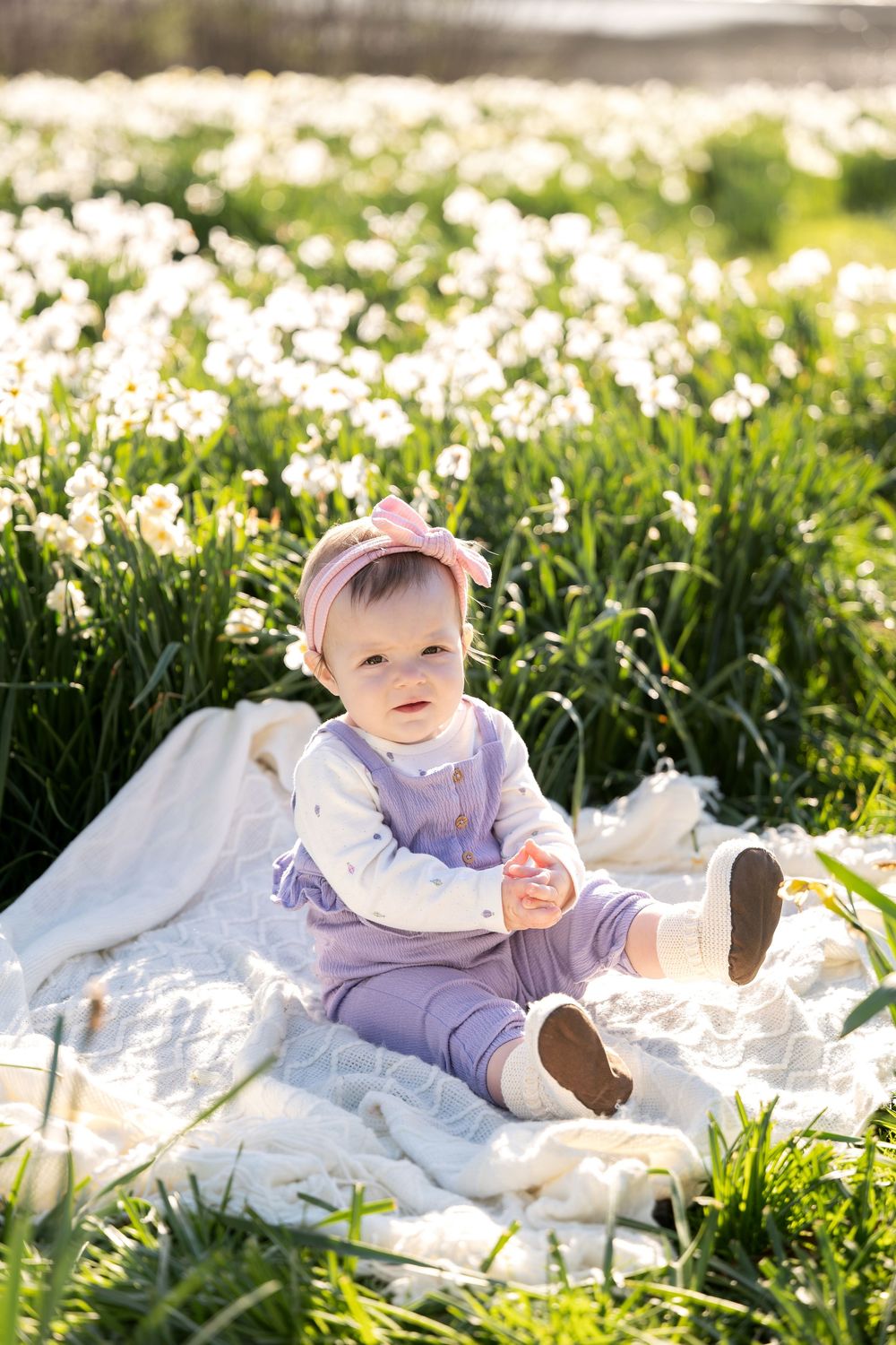 Adorable baby girl in pink bow sitting on blanket surrounded by white daffodils in spring.