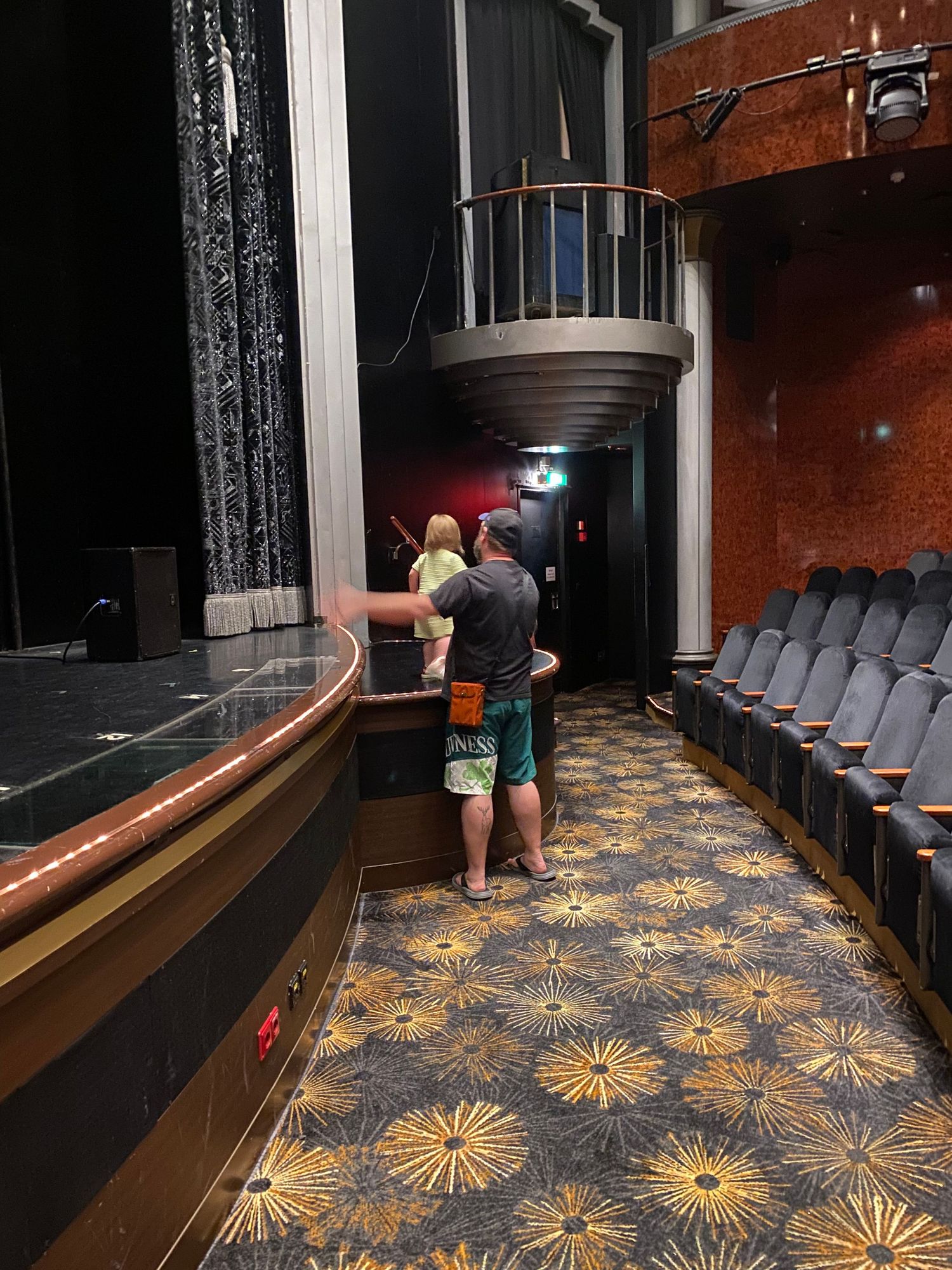 Adults and child explore a theater interior with ornate starburst carpet, stage, and balcony seating area.