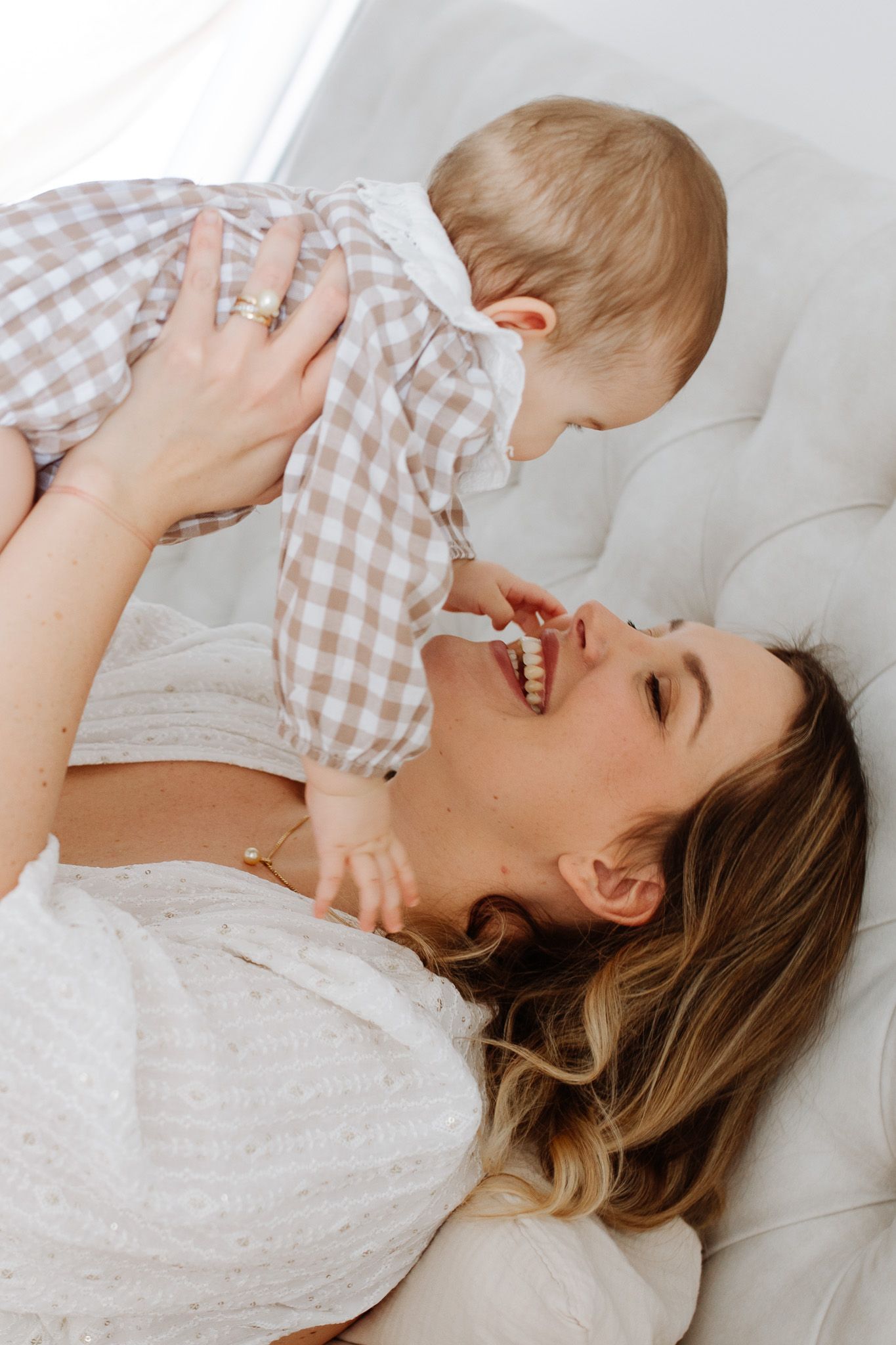 Une mère souriante allongée sur un lit blanc, soulevant joyeusement son bébé vêtu d’une tenue à carreaux
