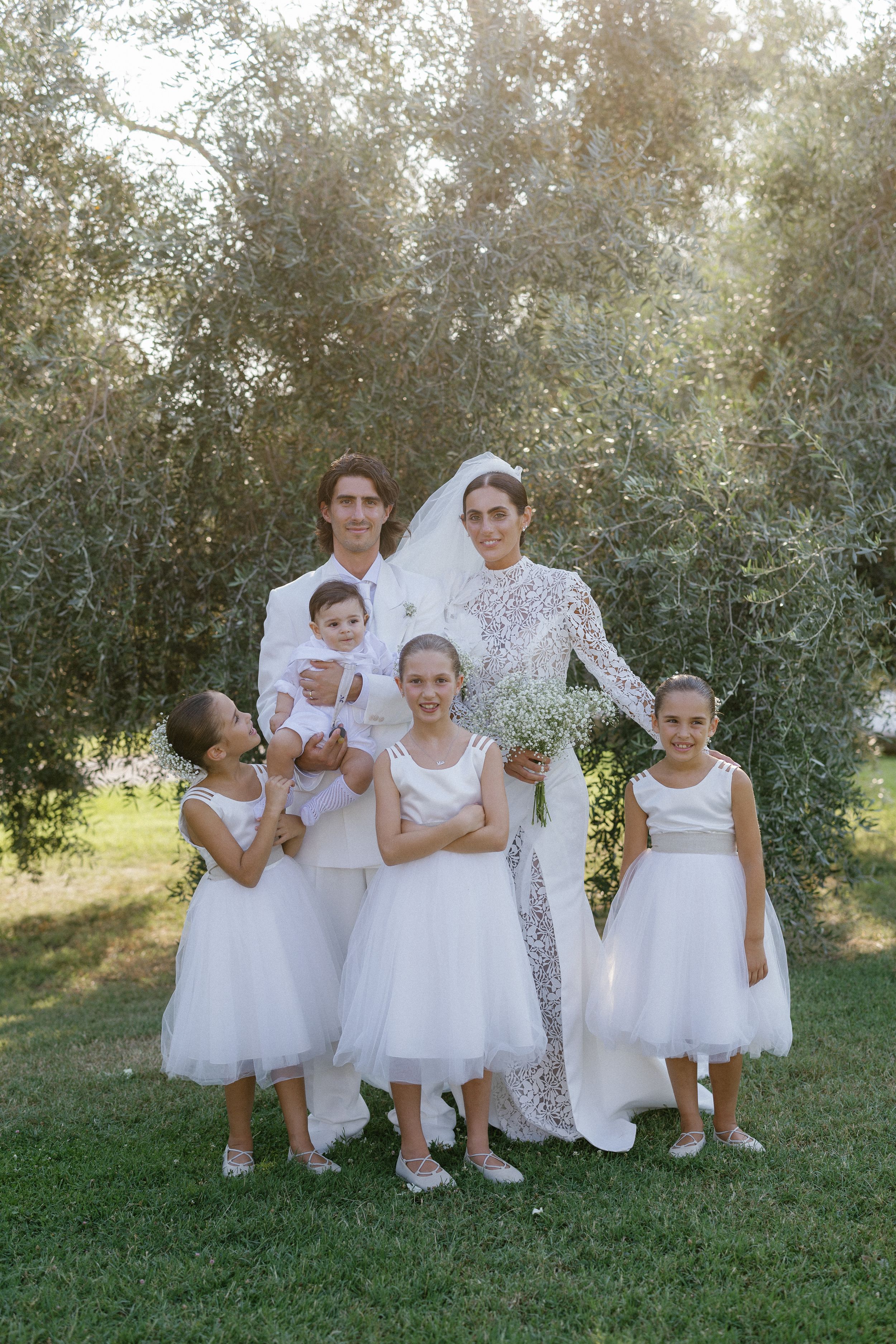 Wedding couple in white with flower girls and baby pose together under olive trees in garden setting.
