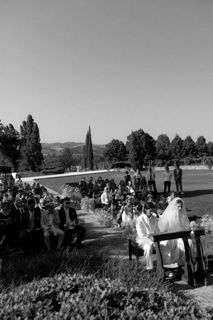 Black and white outdoor wedding ceremony with guests seated along a garden aisle, rolling hills and cypress trees in background.