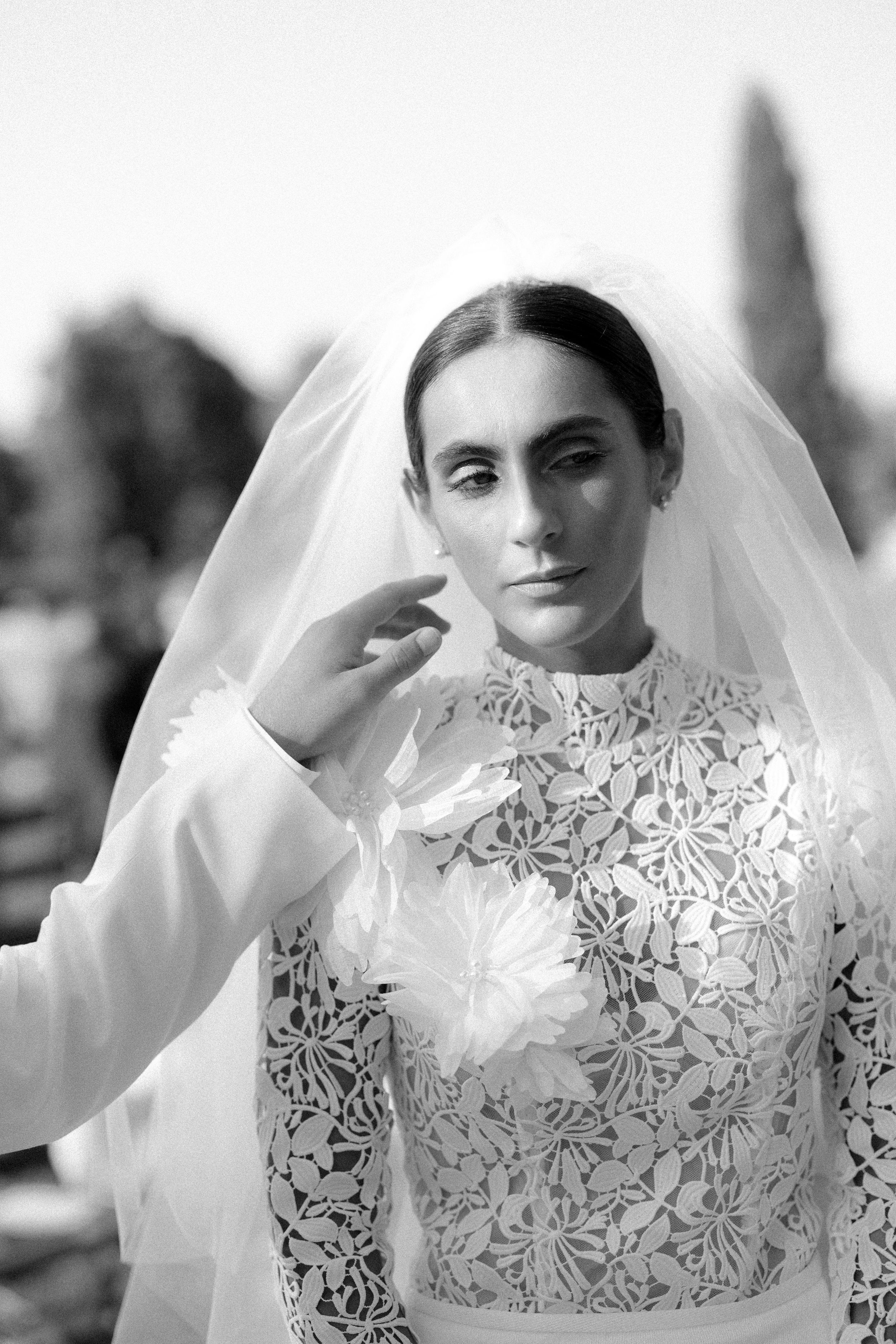 Black and white portrait of a bride in lace high-neck gown adjusting her flowing veil outdoors.