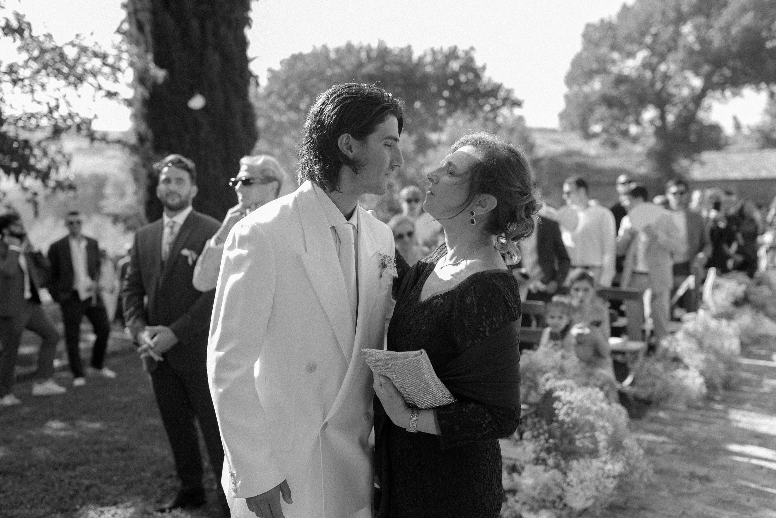 Groom in white suit shares tender moment with guest at outdoor wedding ceremony.