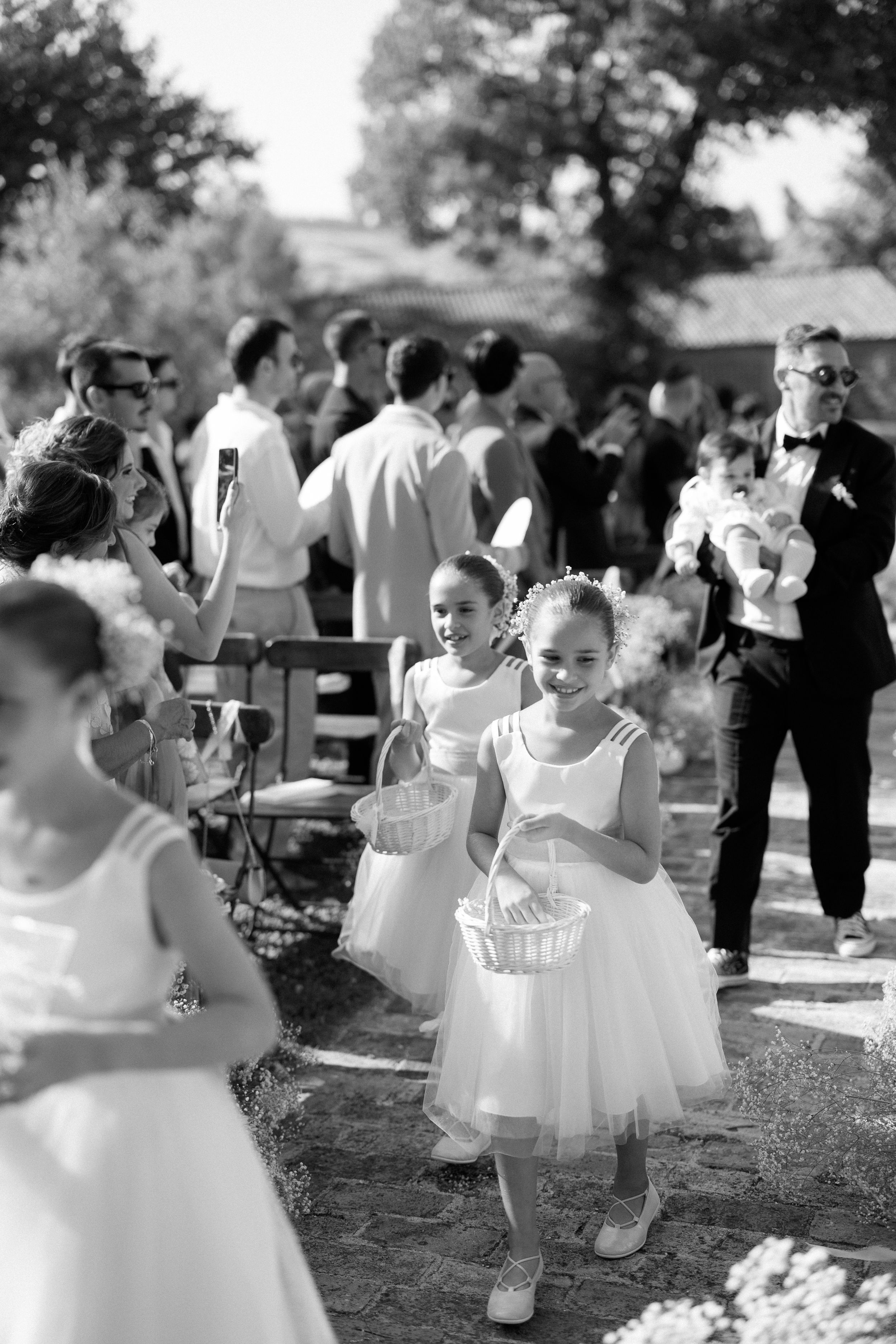 Flower girls in white dresses carry baskets down the aisle at an outdoor wedding ceremony in black and white.