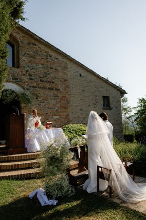 Bride in long veil kneels before priest at rustic stone chapel outdoor wedding.