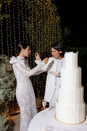 Bride and groom feed each other cake at elegant outdoor wedding with fairy light backdrop.