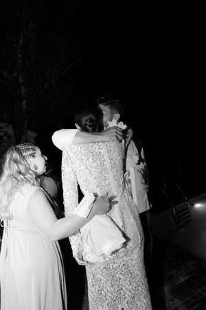 Two brides share an emotional hug during their wedding reception at night, with a bridesmaid nearby.