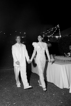 Bride and groom in white wedding attire hold hands walking at outdoor evening reception with string lights.