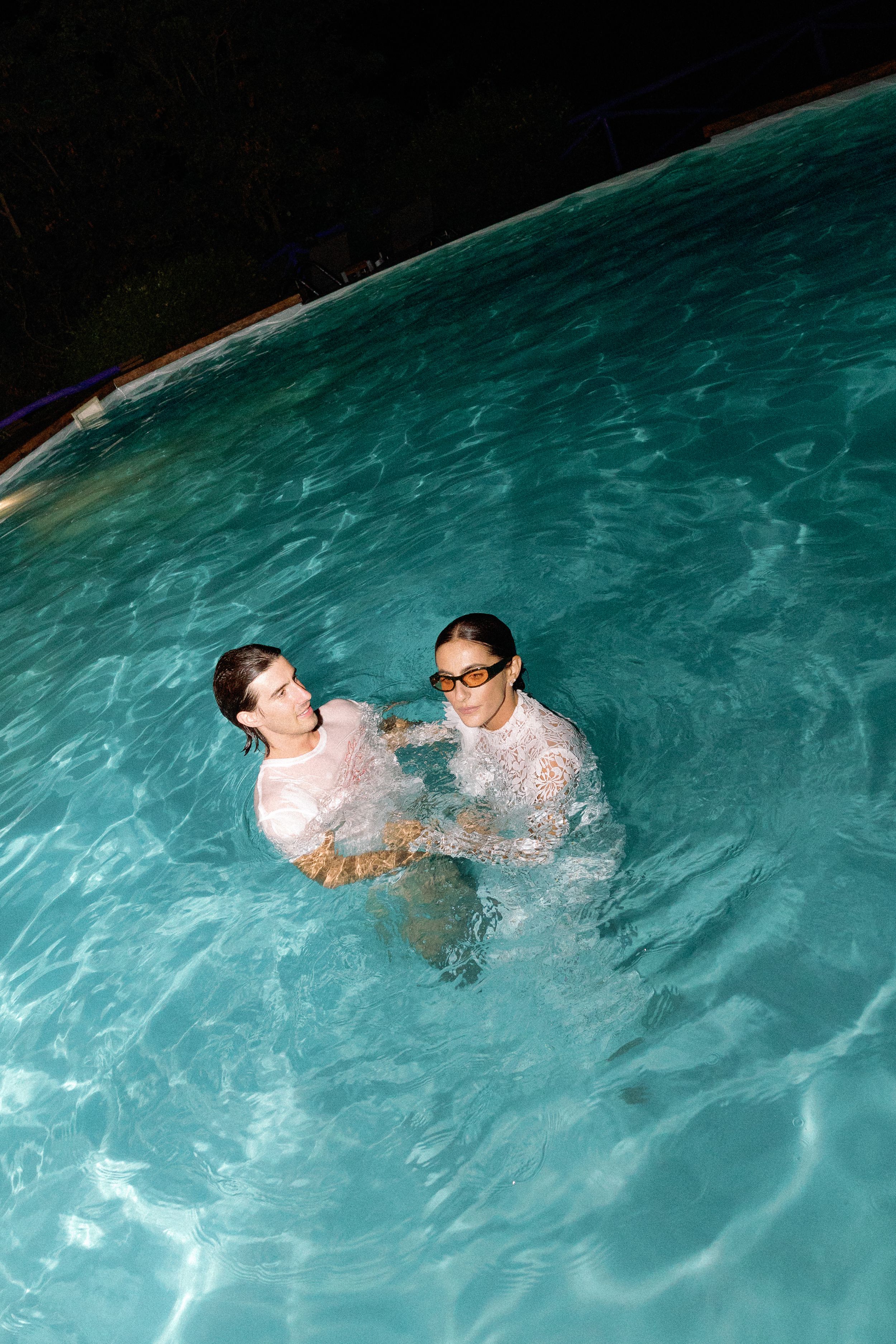 Bride and groom in wedding attire swim together in illuminated pool at night.