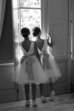 Two flower girls in white tulle dresses with large bows look out a sunlit window.