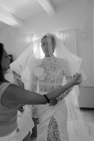 Bride in lace gown gets veil adjusted by assistant before wedding ceremony.