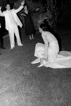 Bride in lace wedding gown kneeling on grass while guests celebrate at outdoor evening wedding reception.