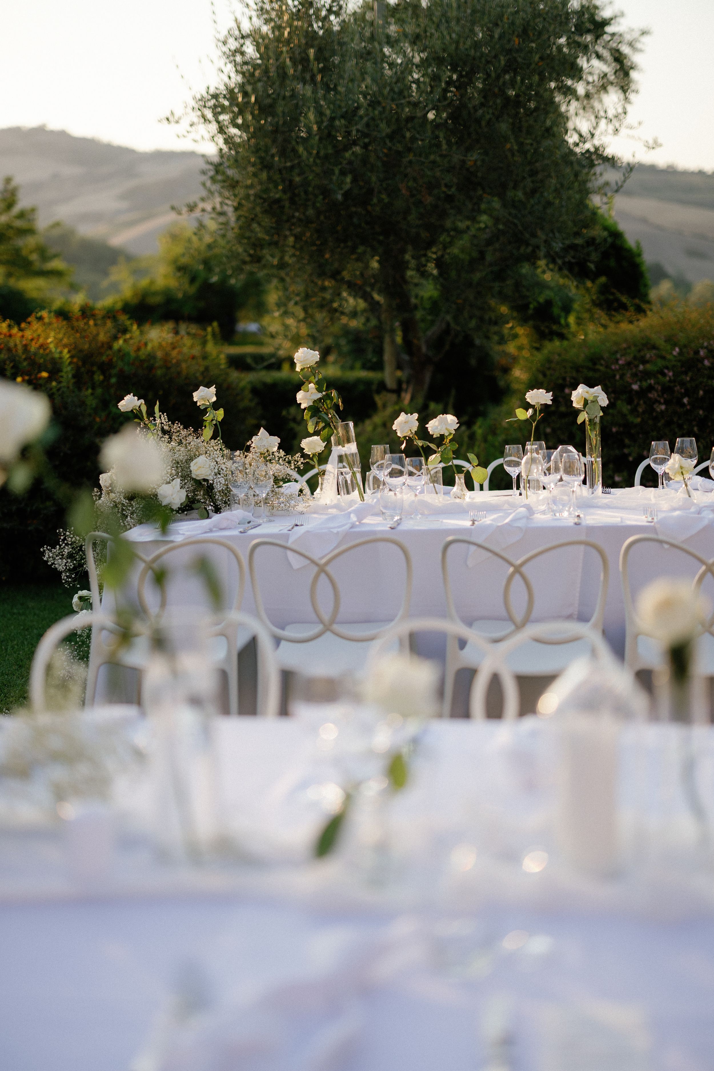 Elegant outdoor wedding reception table with white floral arrangements, glassware, and rolling Tuscan hills in the background.