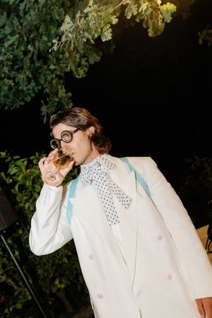 Stylish groom in white double-breasted suit with polka dot bow tie sipping champagne at night.