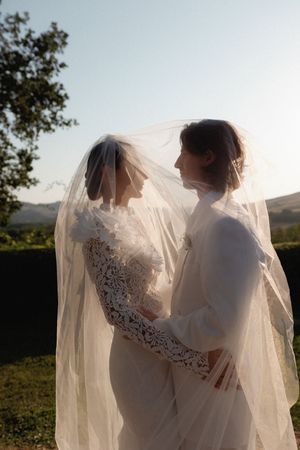 Two brides sharing an intimate moment under a flowing veil at golden hour in a scenic landscape.