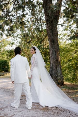 Bride and groom in white attire walk hand in hand on a gravel path surrounded by lush green trees.