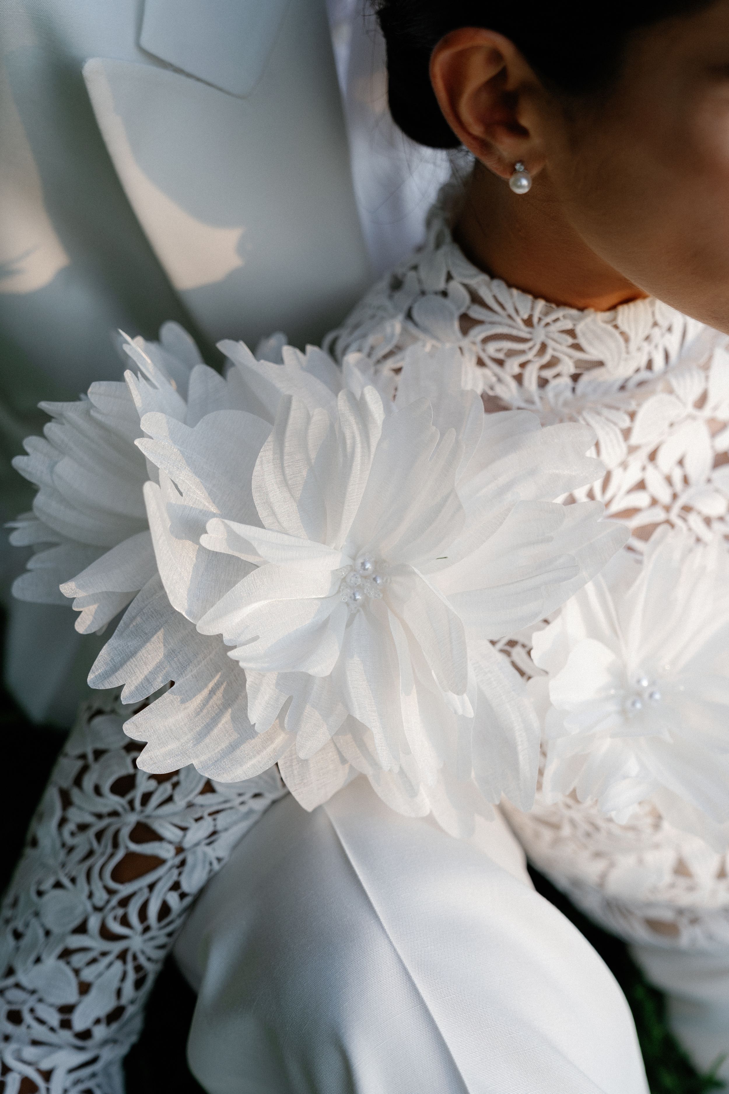 Close-up of bride wearing intricate lace gown with 3D floral applique and pearl earring on wedding day.