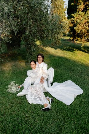 Couple in white wedding attire sitting on green grass surrounded by lush trees in a sunlit garden.