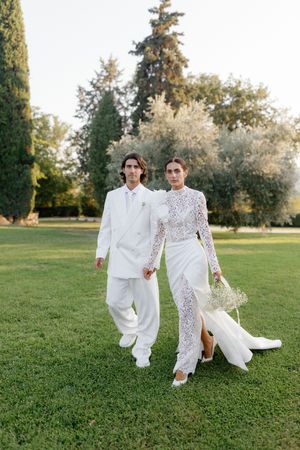 Bride and groom in white wedding attire holding hands while walking through lush garden grounds.