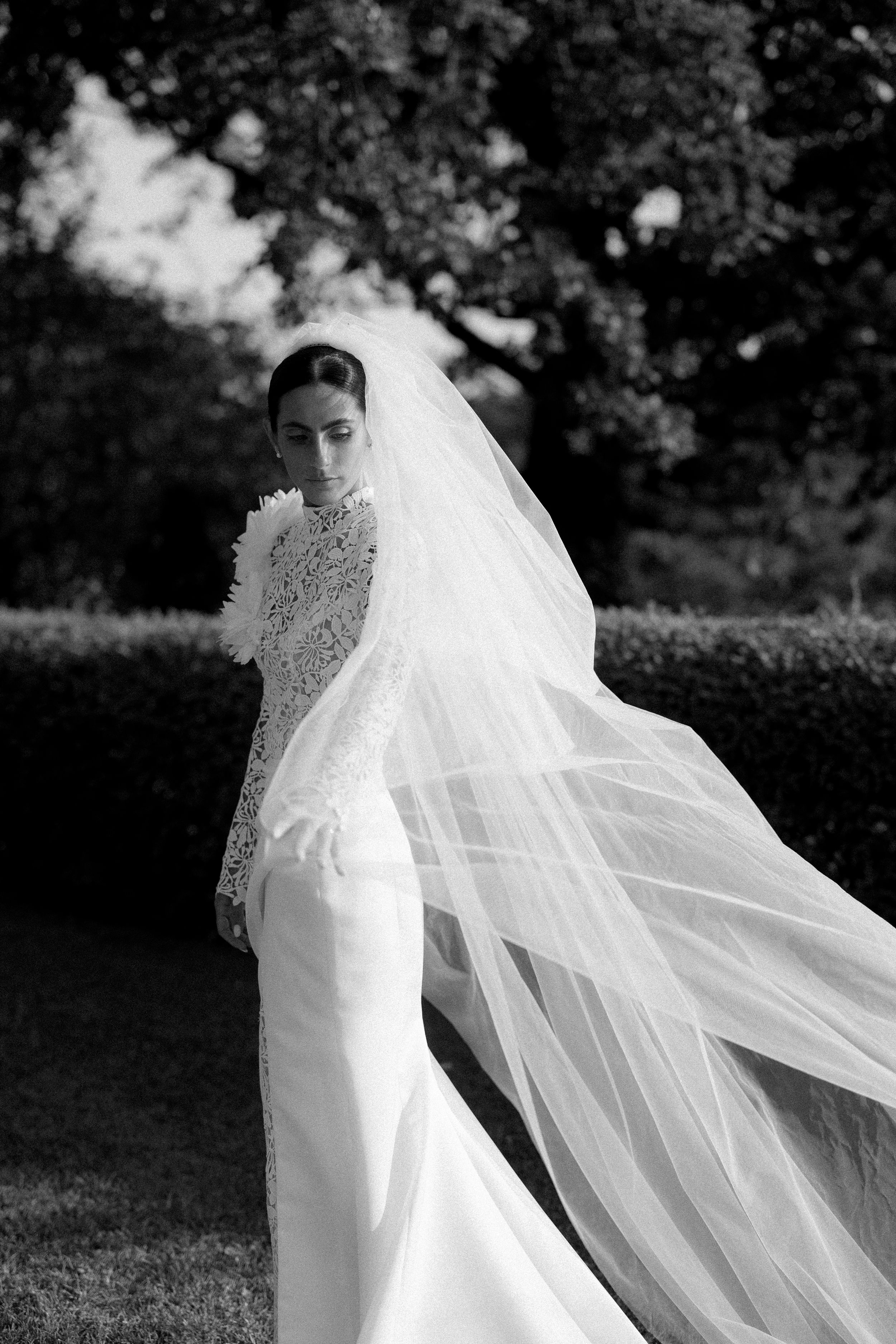 Black and white photo of a bride in lace gown with dramatic flowing veil billowing in a garden.