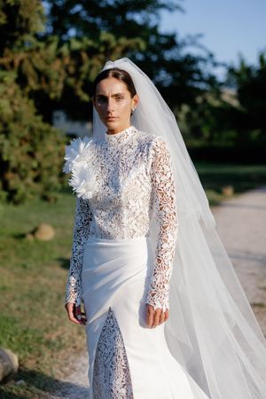 Bride in white lace high-neck gown with long veil and floral detail, posing outdoors among green trees.