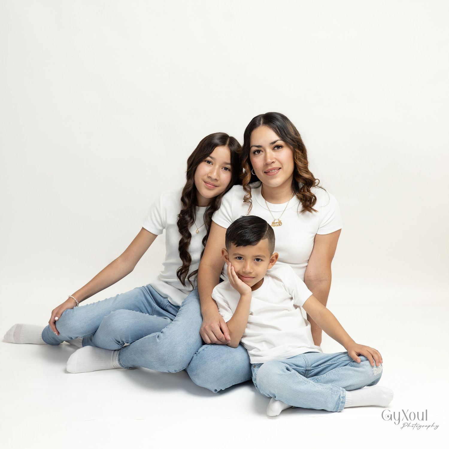 A mother and two children pose together in matching white tops and jeans against a white studio background.