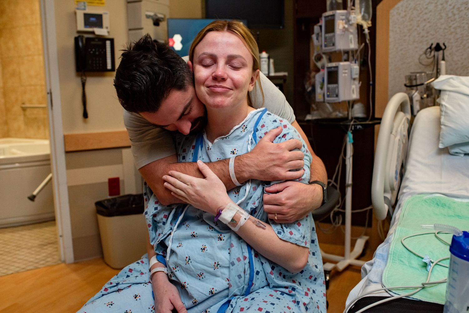 A woman in a hospital gown receives a comforting hug from a man in a medical room setting.