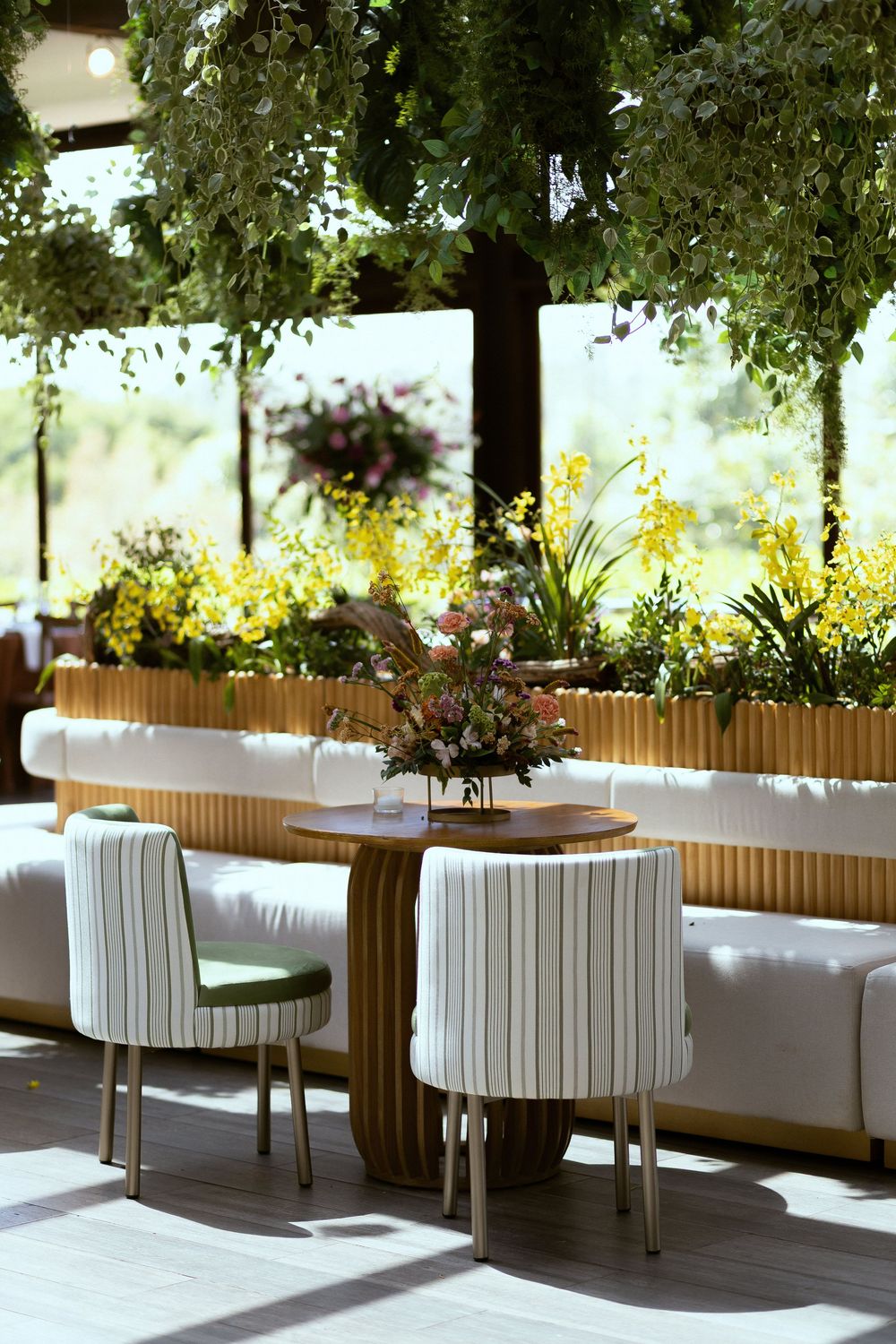 Elegant restaurant seating with white striped chairs, wooden table, fresh flowers, and lush hanging greenery in a sunlit space.