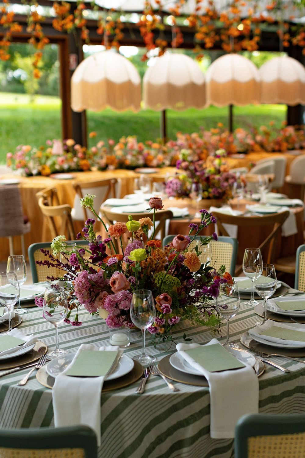 Elegant outdoor wedding reception table with colorful floral centerpiece, striped linens, crystal glasses, and hanging pendant lights.