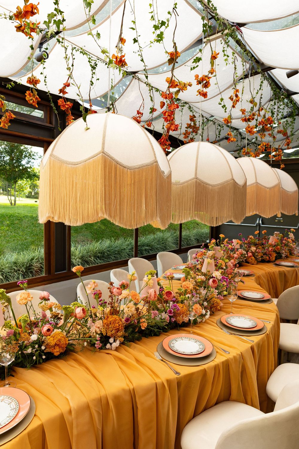 Elegant wedding reception table with gold fringe pendant lights, orange floral garland, and lush floral centerpieces under a glass ceiling.