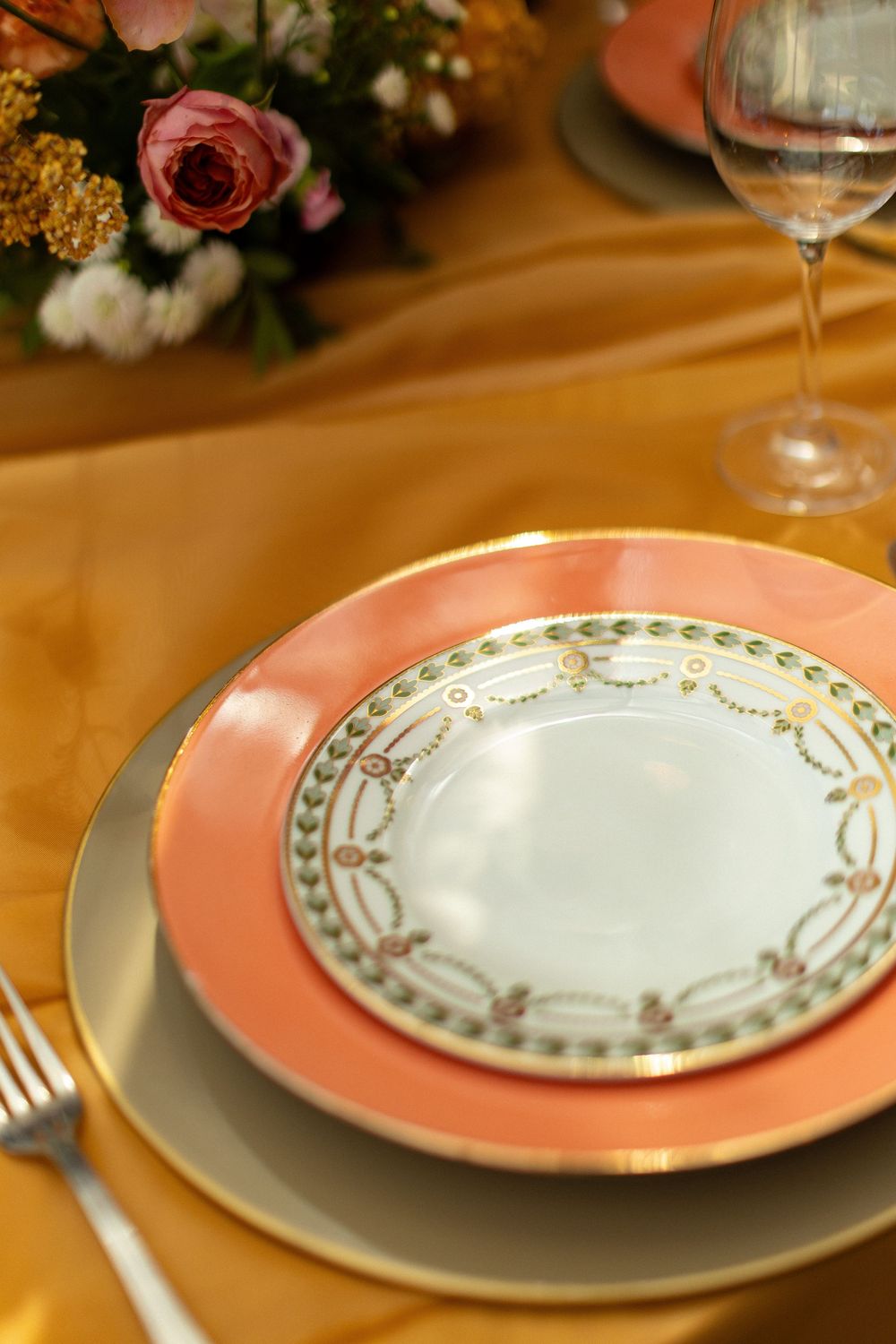 Elegant place setting with pink and gold trimmed china plates on golden satin tablecloth with floral centerpiece.