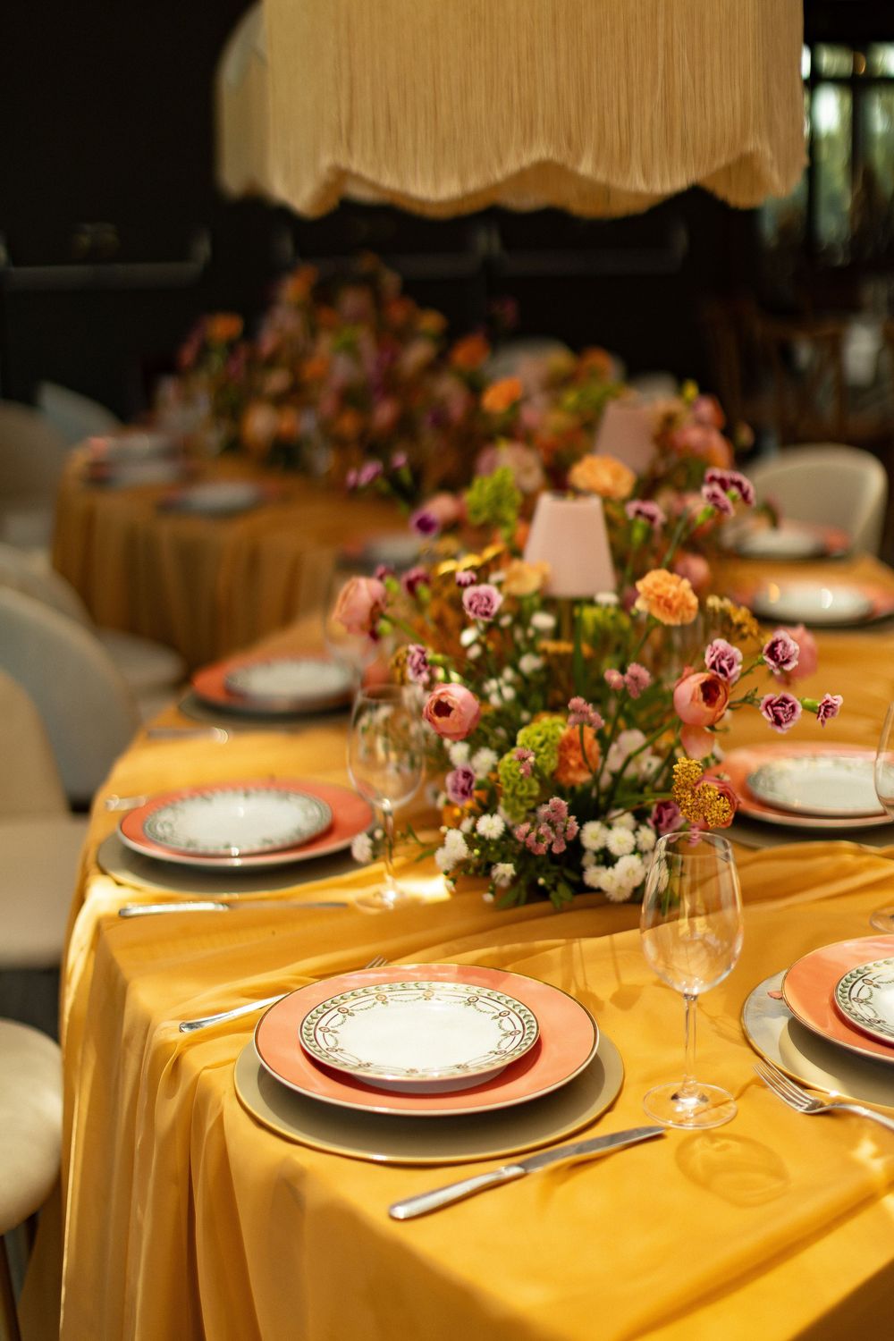 Formal dinner table setting with gold satin tablecloth, decorative plates, crystal glasses, and vibrant mixed floral arrangements.