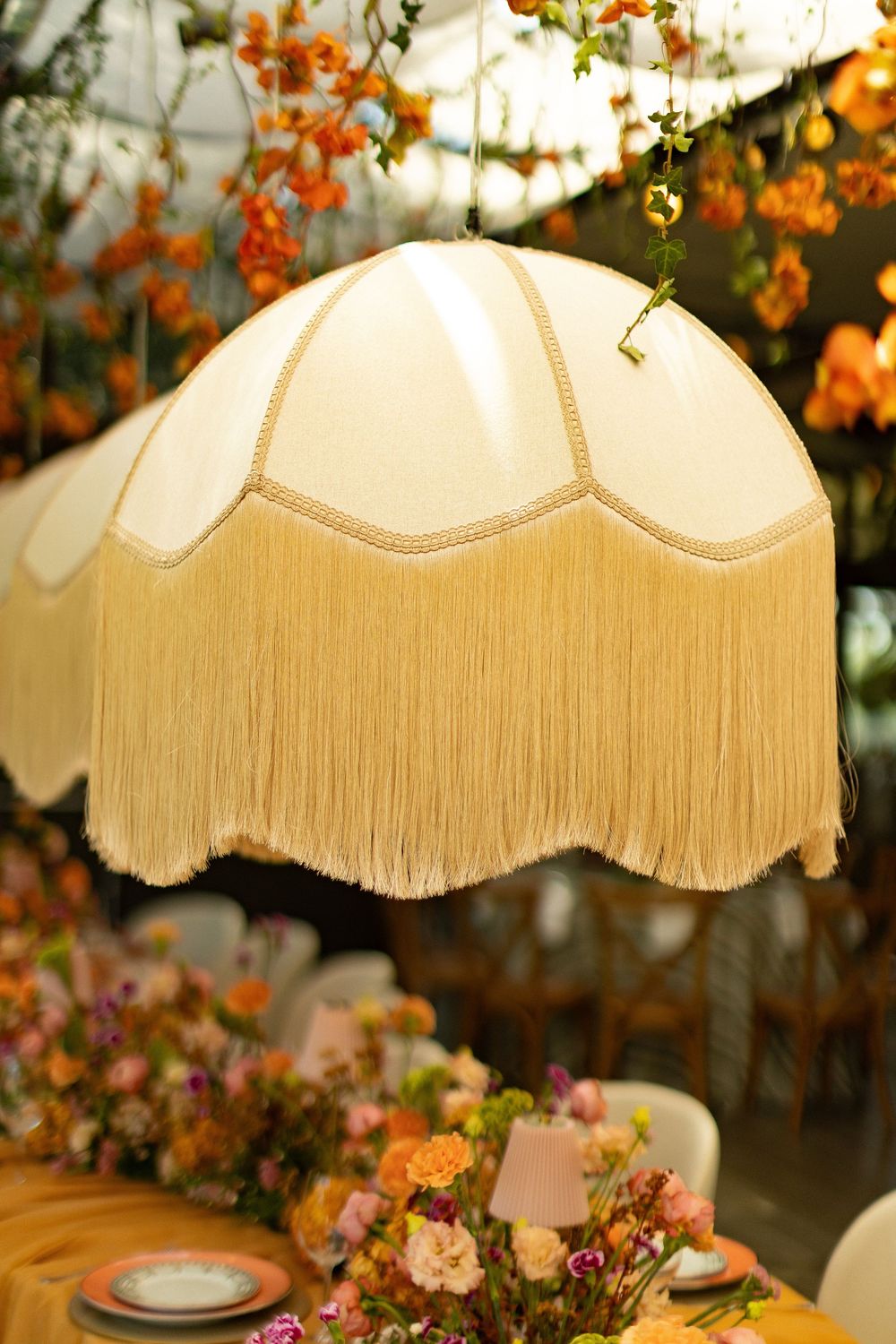 Vintage cream fringed pendant lamp shade hanging over a floral decorated event table with orange and pink flowers.