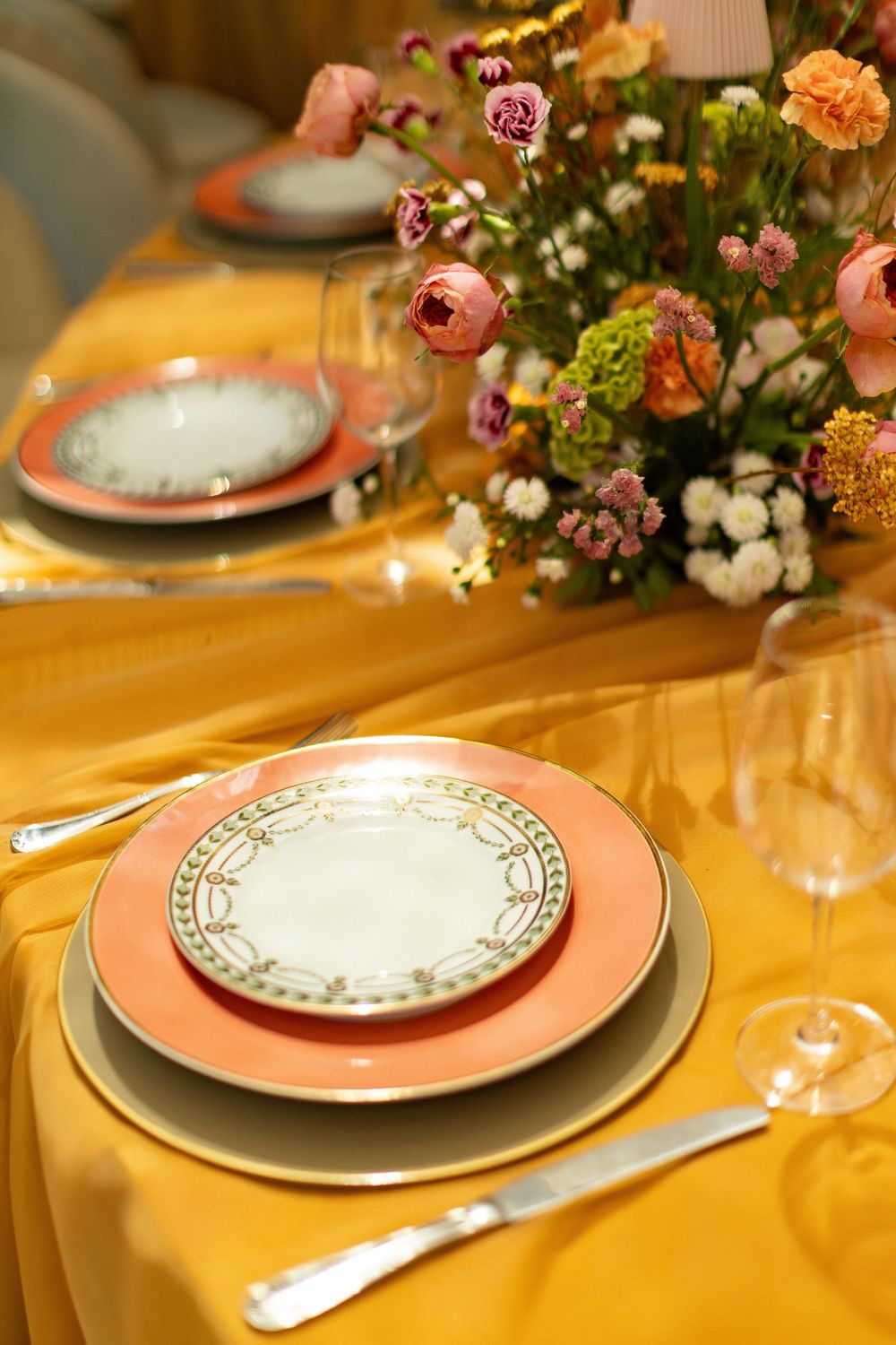 Elegant table setting with gold tablecloth, pink-rimmed plates, crystal glasses, and colorful floral centerpiece.