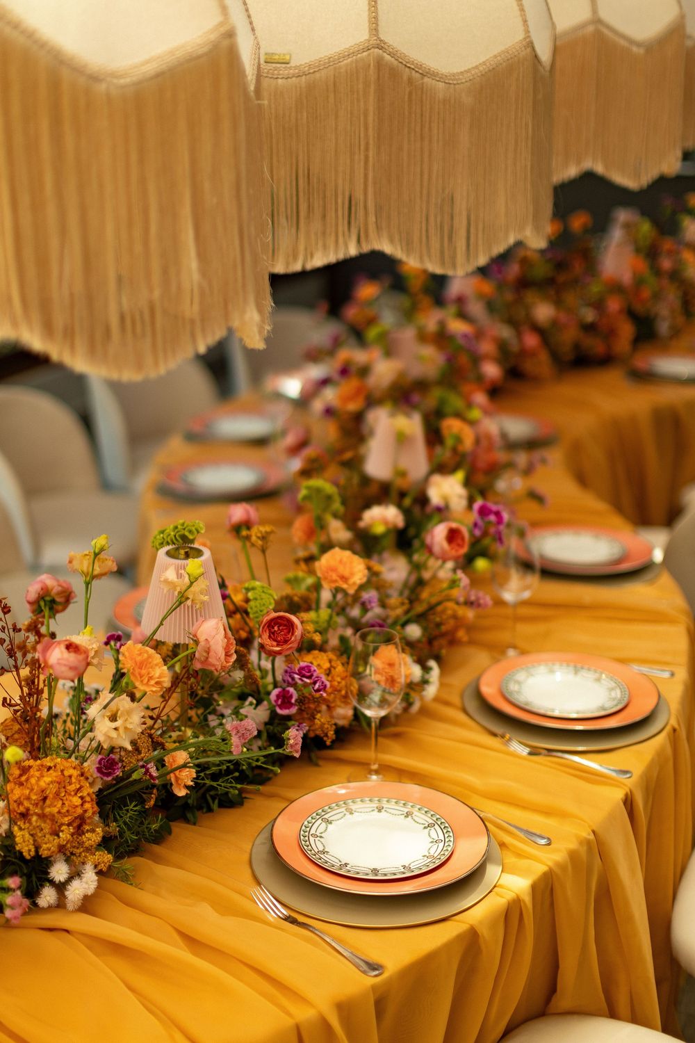 Elegant wedding tablescape with golden yellow linen, colorful floral runner, stacked plates, and fringed pendant lamps overhead.