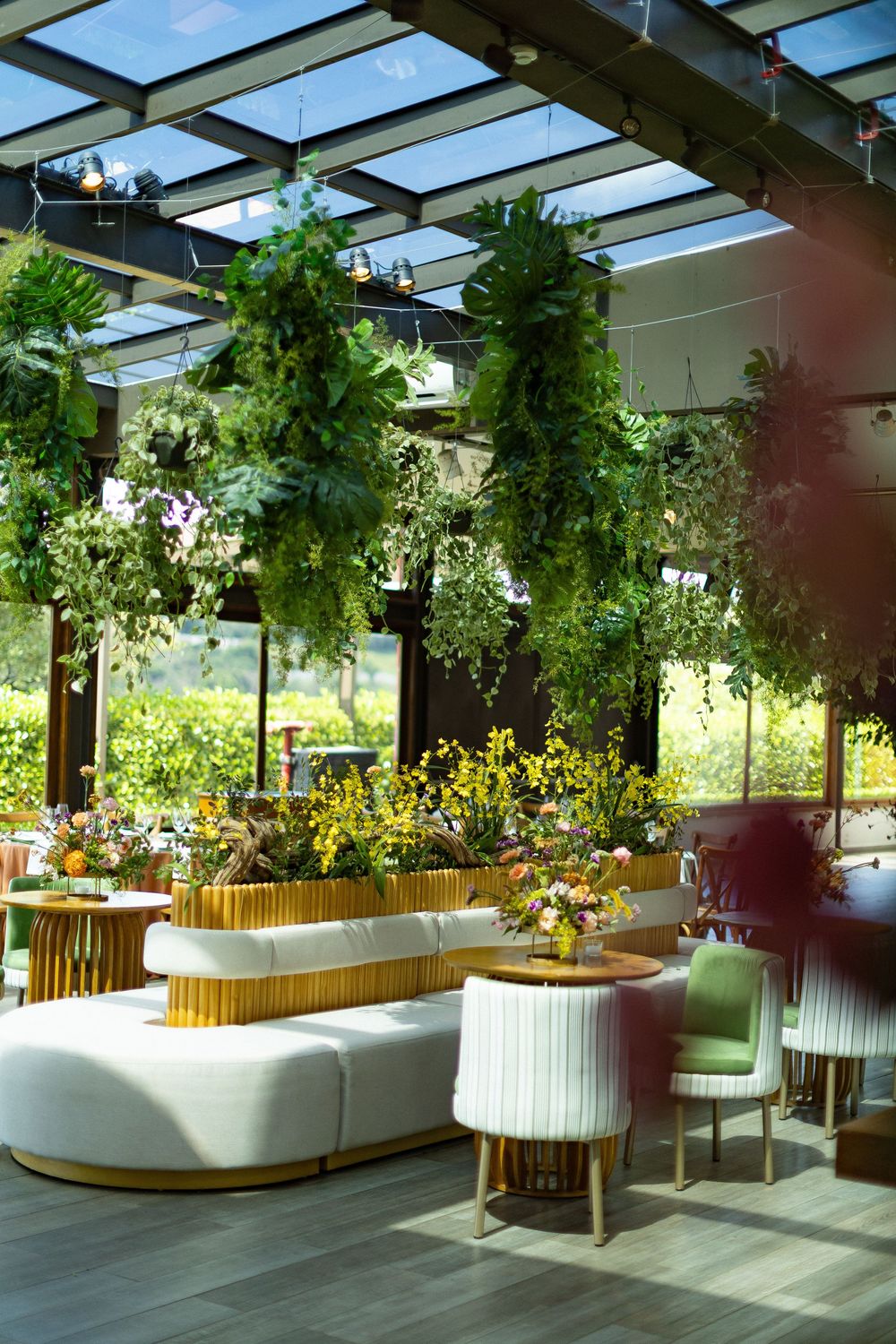 Elegant indoor lounge with hanging greenery, white curved sofa, gold accents, and lush garden views through glass walls.
