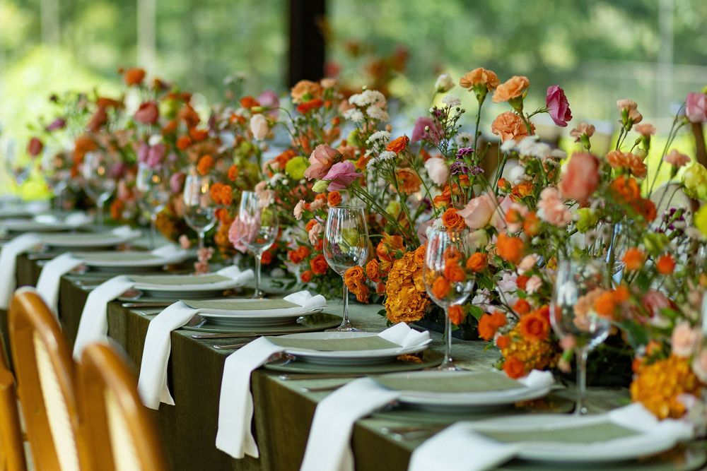 Long banquet table with olive green linen, white napkins, and vibrant orange, pink, and green floral centerpieces.