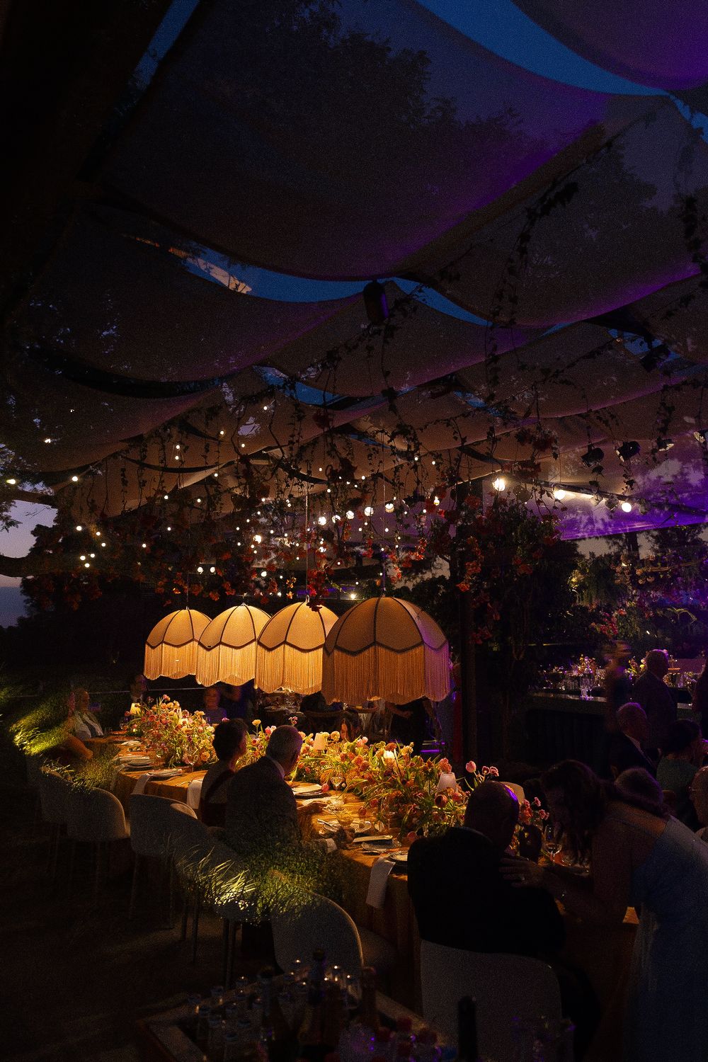 Elegant outdoor wedding reception under a tent with warm glowing lanterns, string lights, and floral centerpieces at dusk.