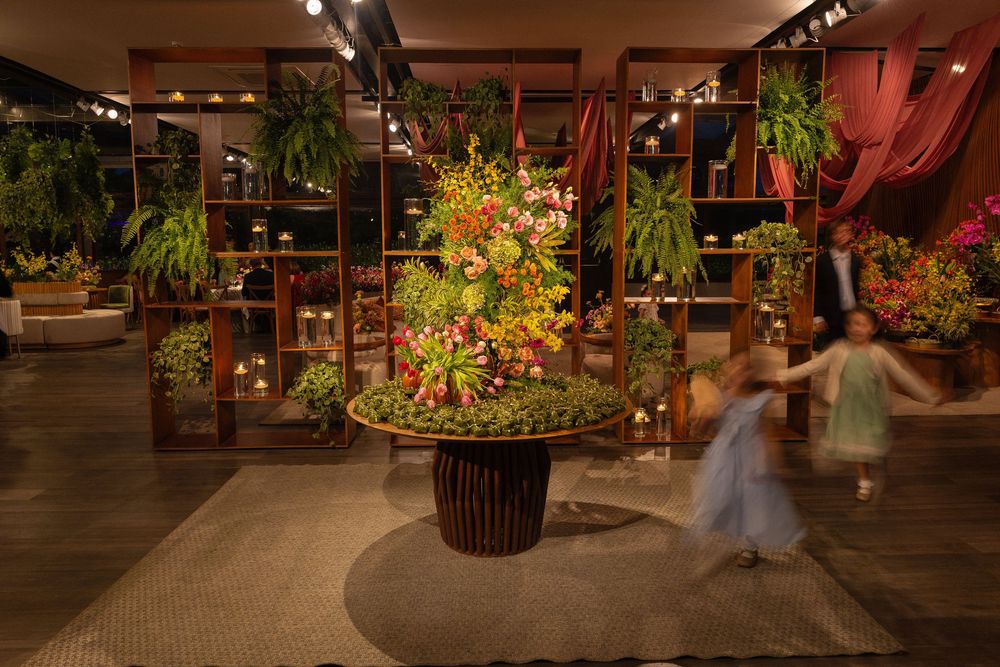 Lush floral tower centerpiece with tropical blooms displayed in a warmly lit indoor space with wooden shelving and blurred figures.