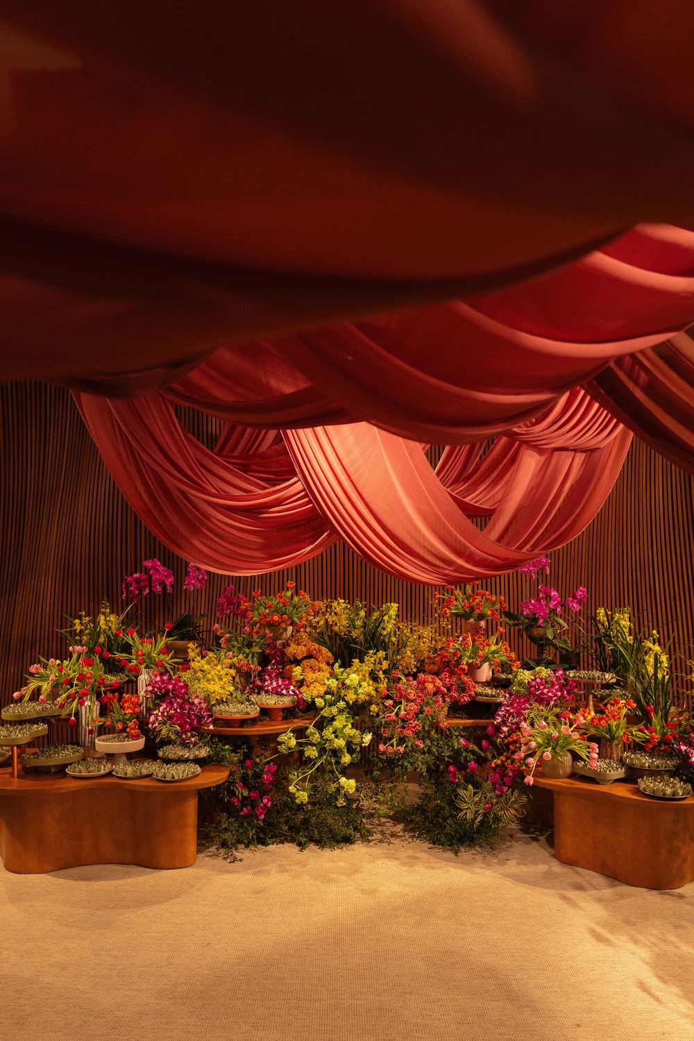 Luxurious event space with draped red fabric ceiling and vibrant floral arrangements on wooden tables in warm lighting.
