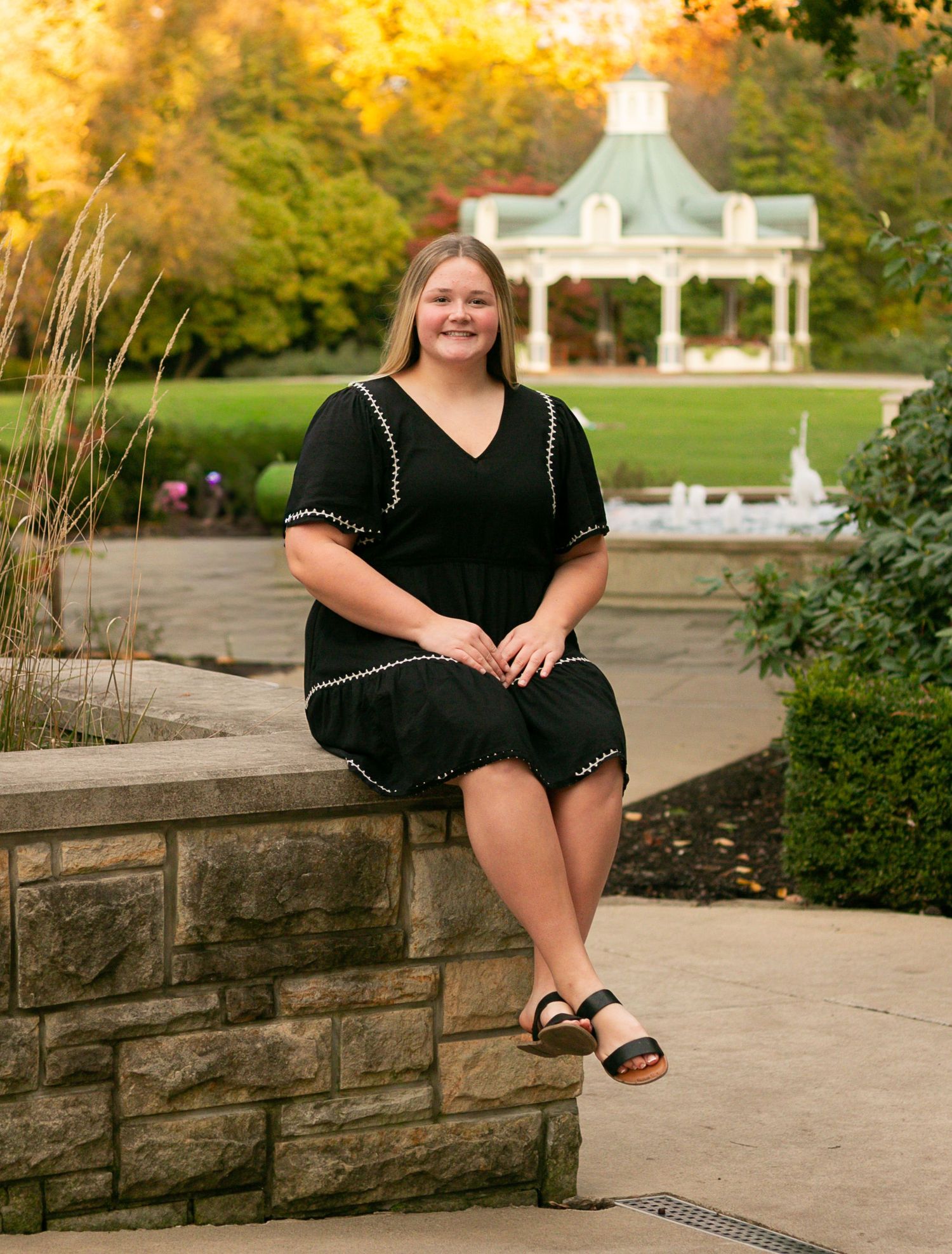 Young woman in black dress sitting on stone wall in park with gazebo and autumn foliage background. mill creek park