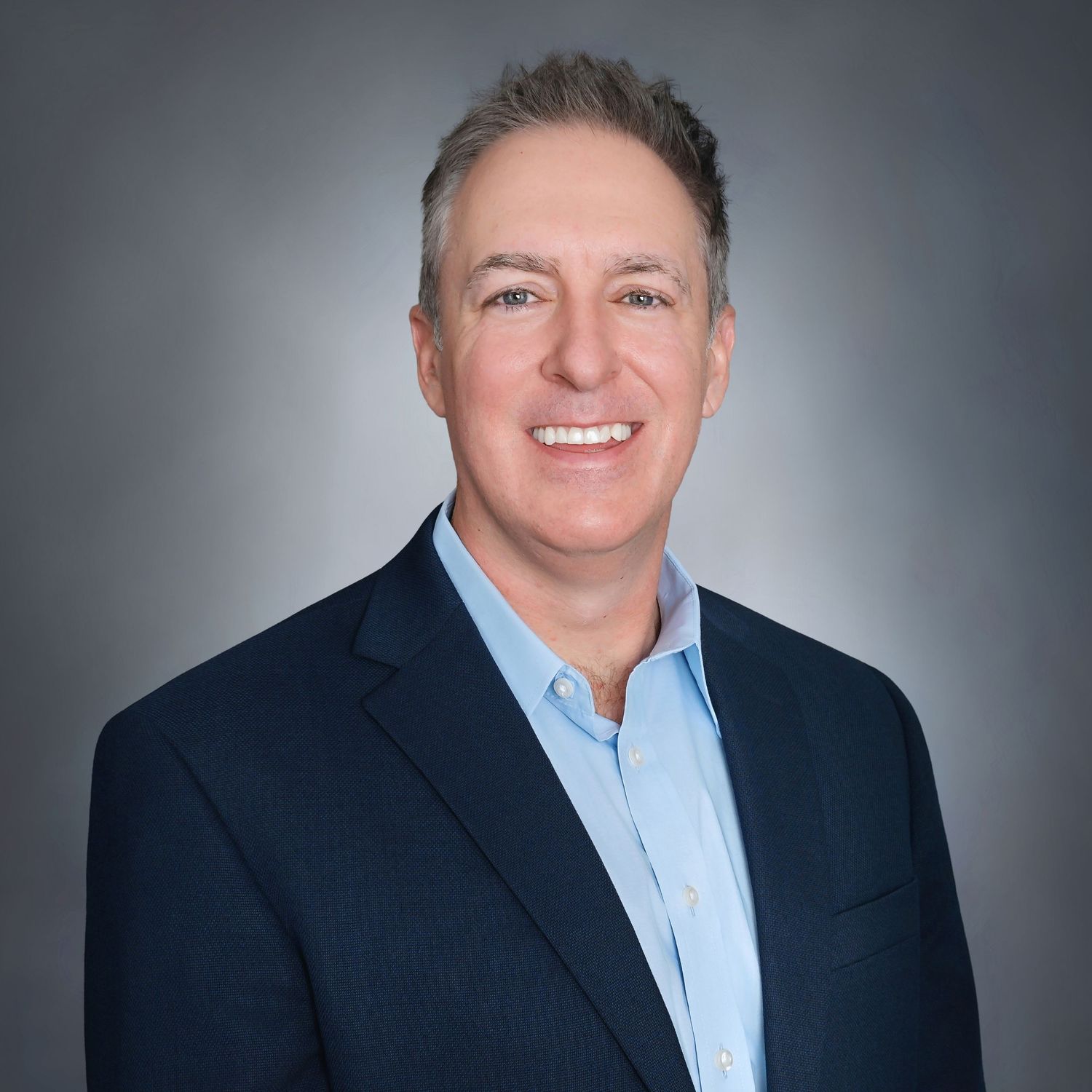 Professional headshot of a smiling middle-aged man in a navy blazer and light blue shirt against a gray background.
