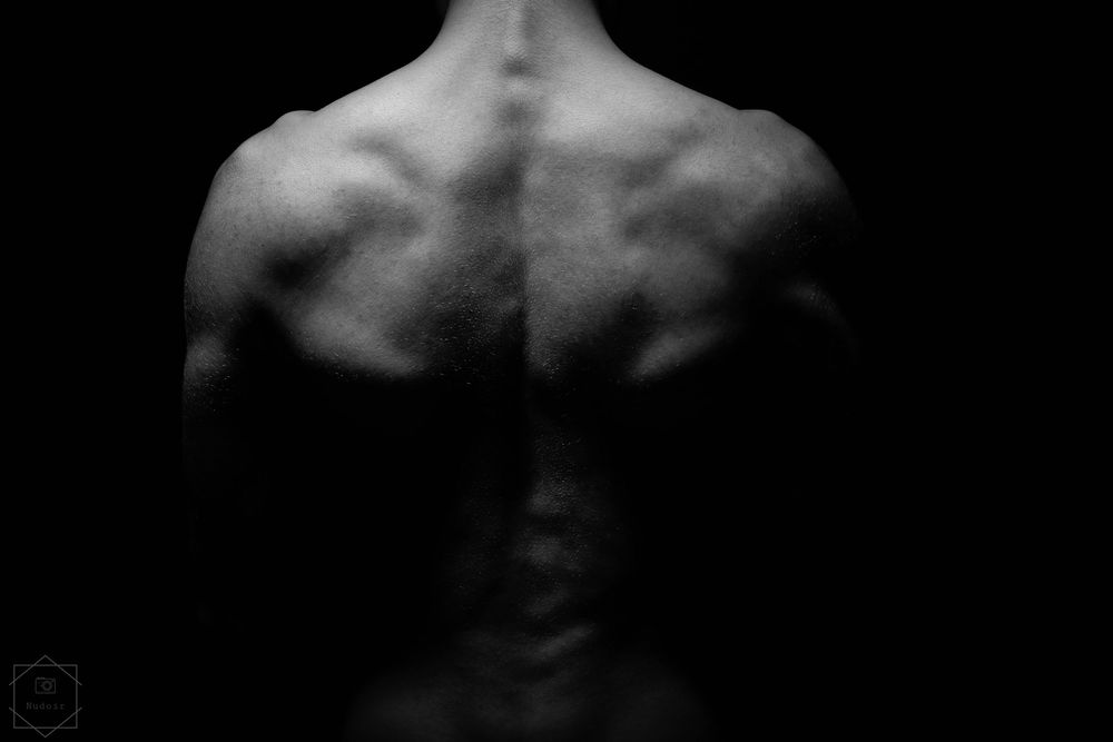 Black and white photo of a muscular male back showing defined muscles and spine in dramatic low-key lighting.