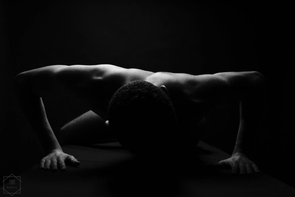 Black and white artistic photo of a shirtless man in a low push-up pose against a dark background.