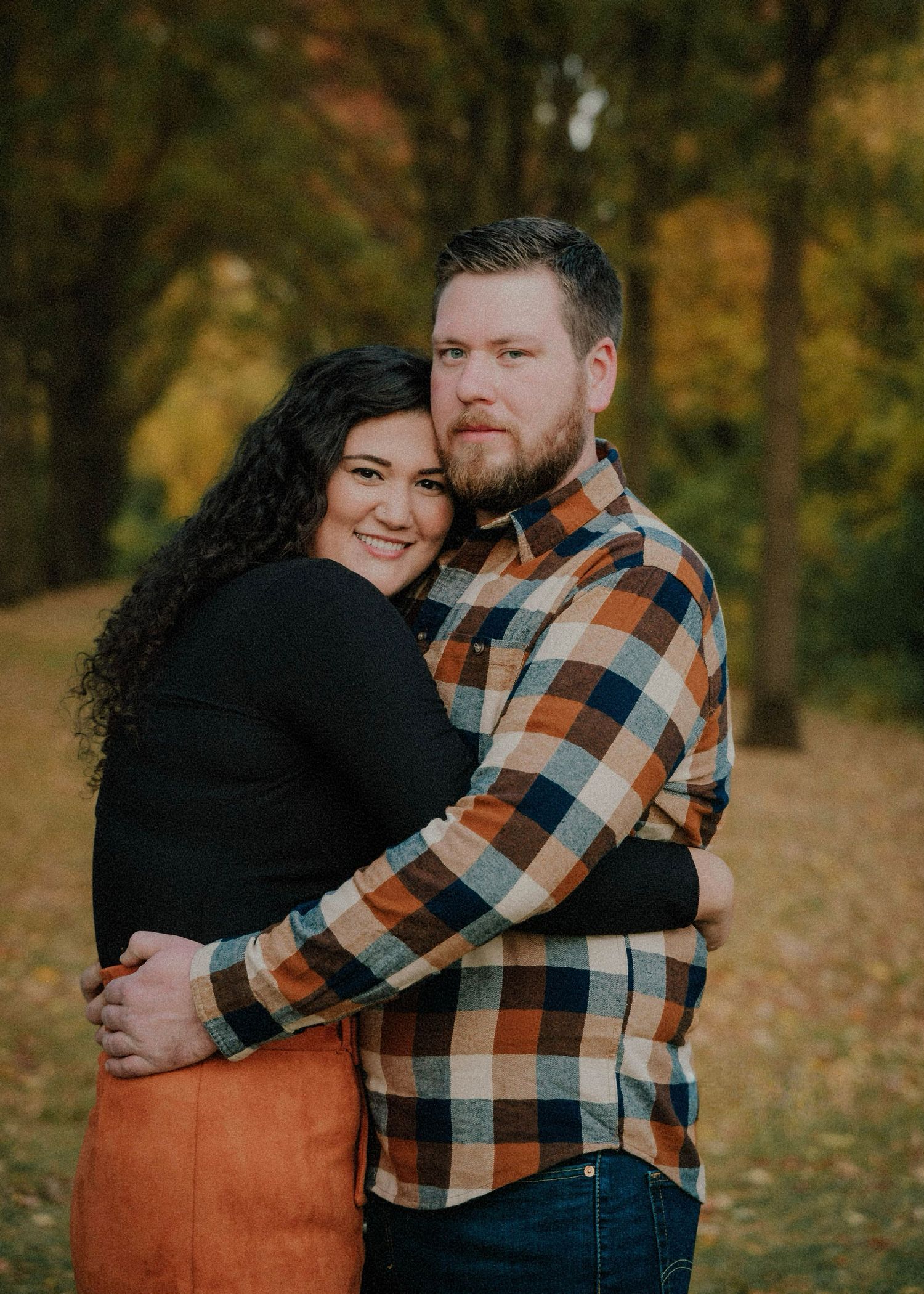 Couple embracing in autumn park, woman in black top and orange skirt, man in plaid flannel shirt, fall foliage background.